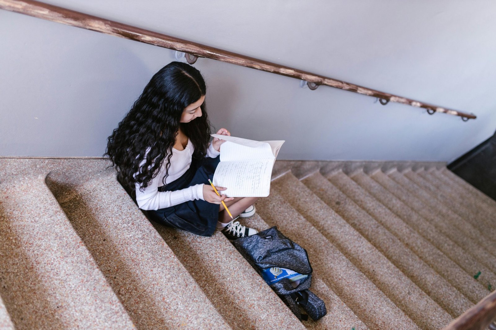 A young girl studies on concrete stairs with a backpack and notebook in a school hallway.